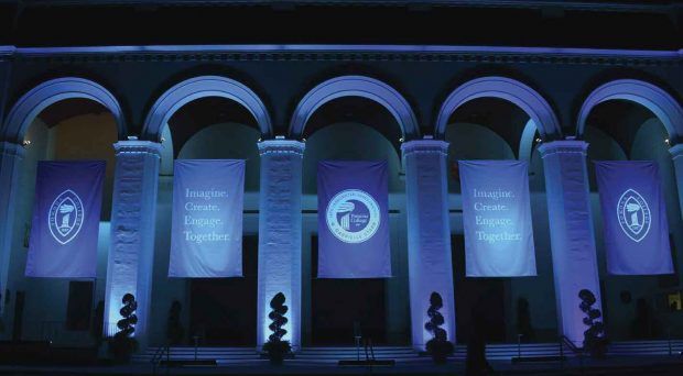 Dramatic lighting on the front of Bridges Auditorium reveals banners with the College mark, the inaugural logo and the theme of the inauguration—”Imagine. Create. Engage. Together.”